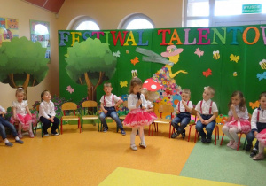 Dziewczynka trzyma w ręku mikrofon, recytuje wiersz. Wokół dziewczynki siedzą dzieci.